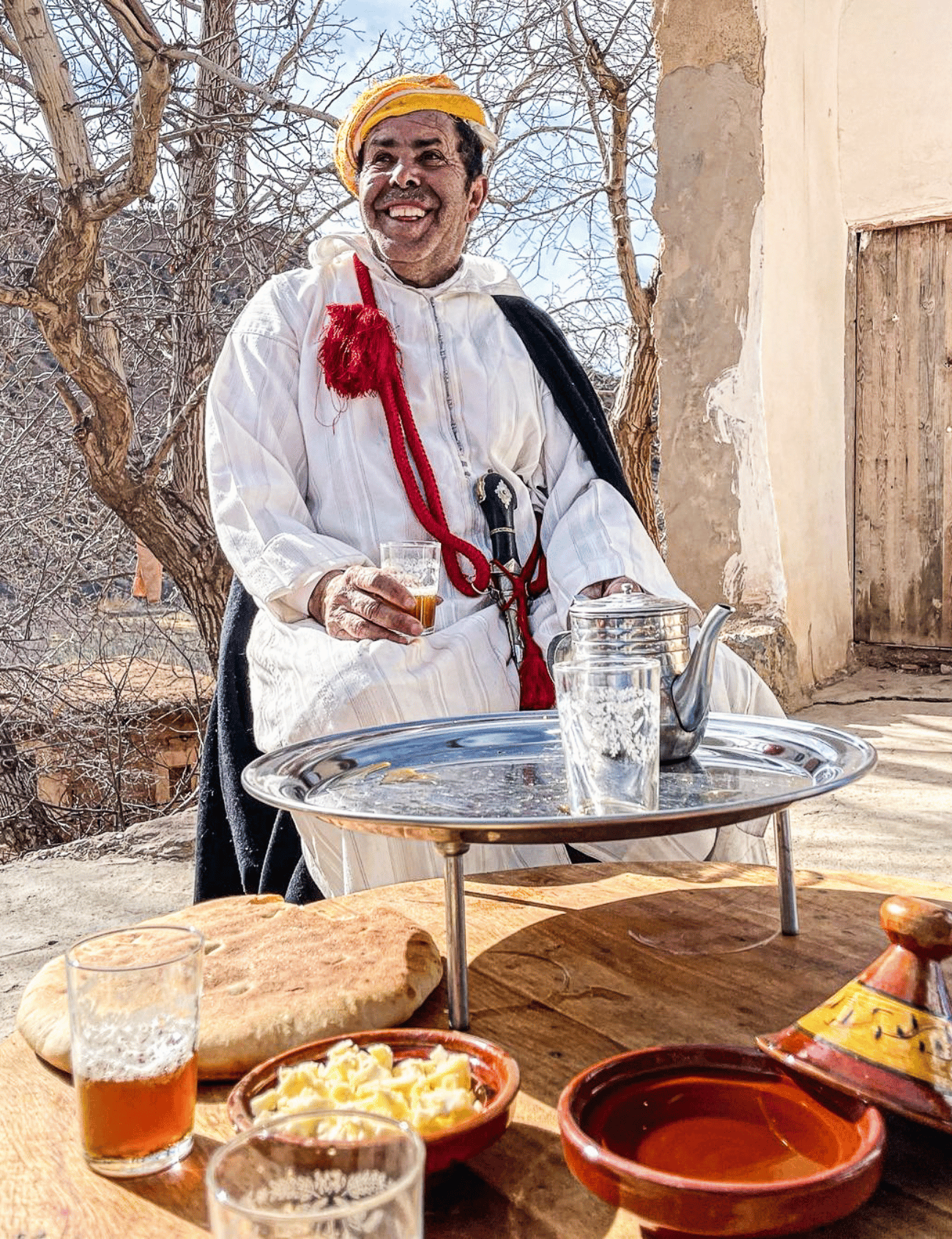 Smiling Moroccan man in traditional dress serving tea outdoors, with fresh bread and butter on the table.